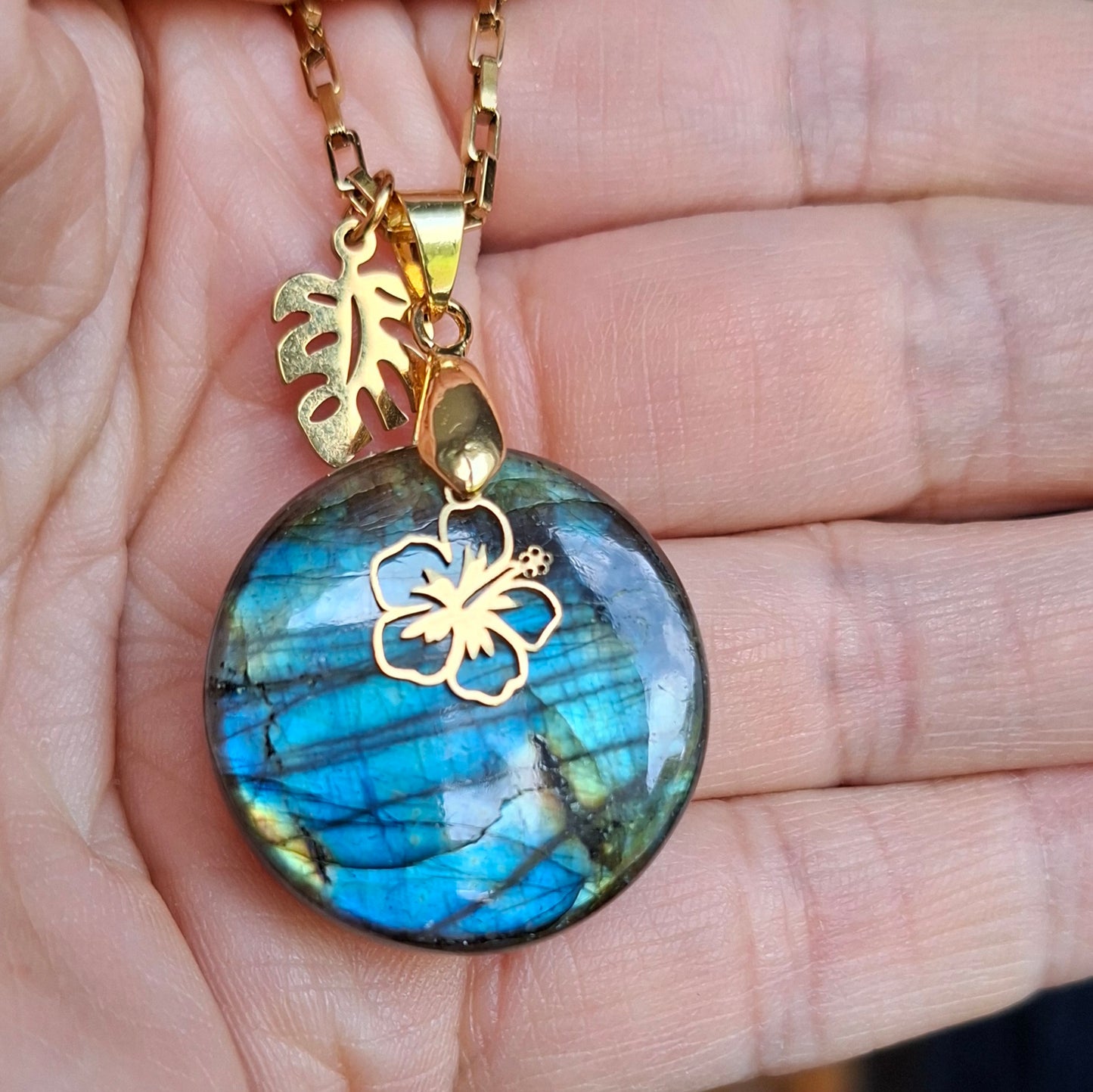 Collier en labradorite ronde présenté dans la main avec fleur d'hibiscus et feuille dorées.