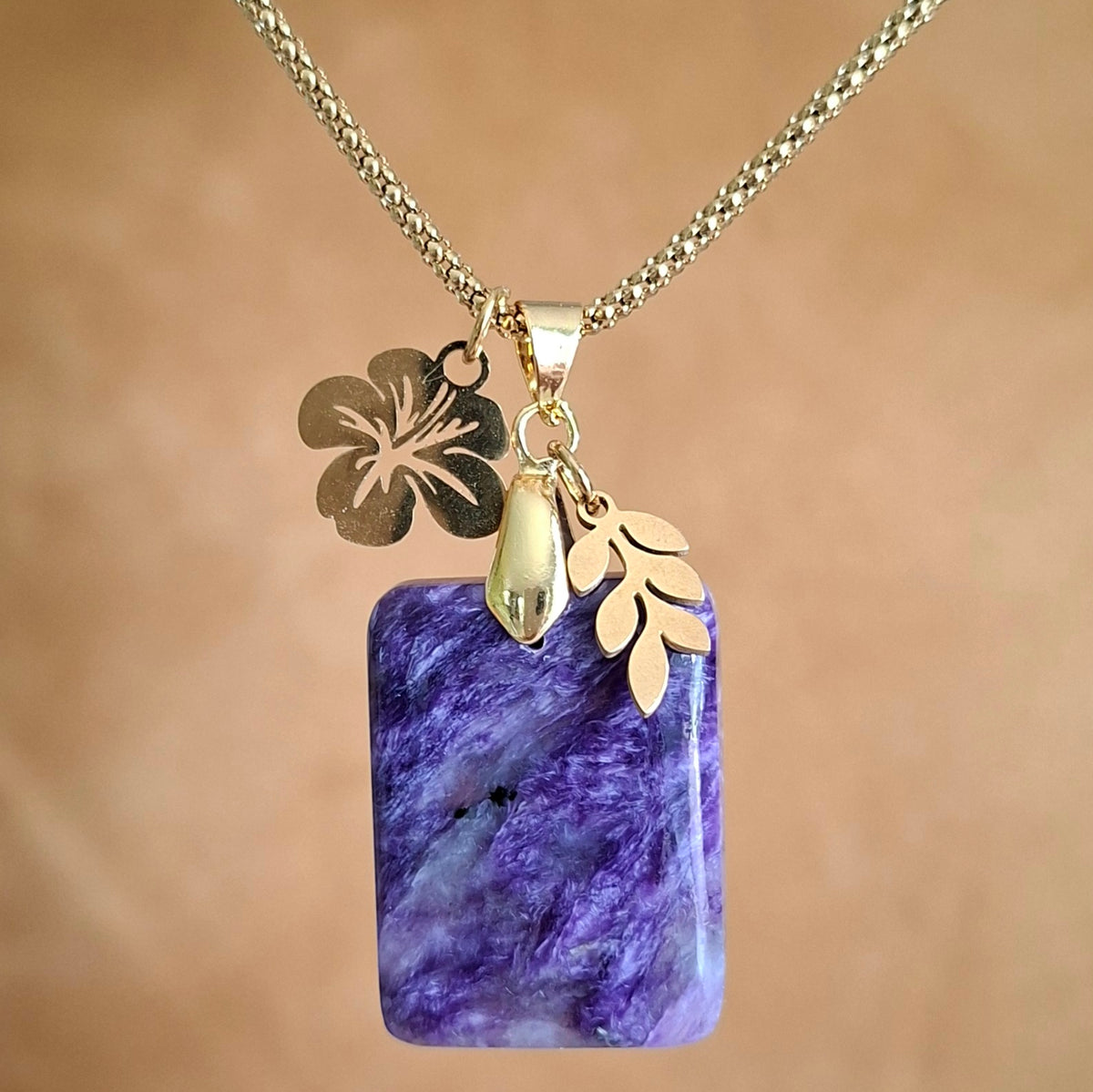 Collier en charoïte rectangulaire violette avec breloque feuille et fleur d'hibiscus dorées.