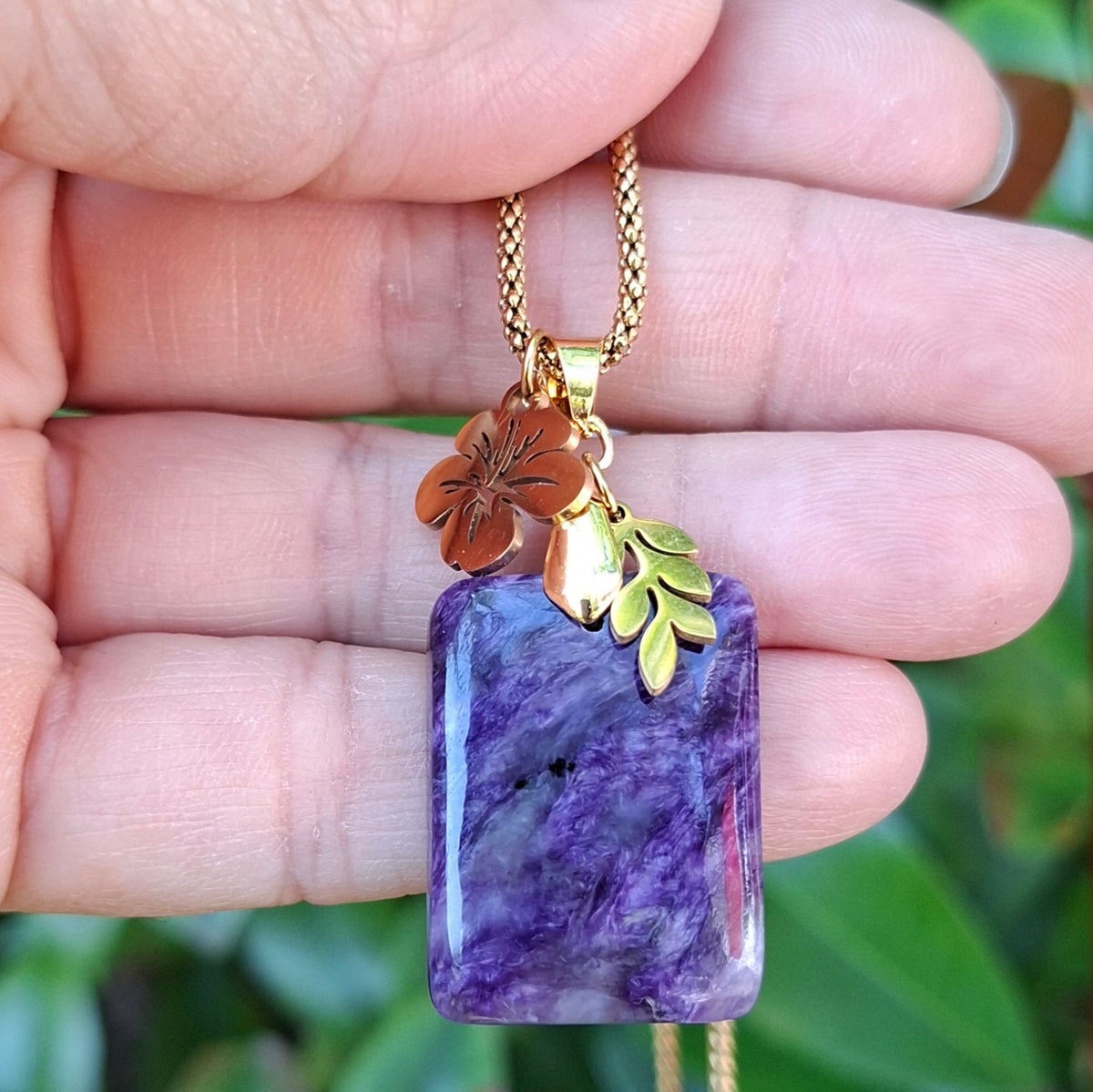 Collier en charoïte rectangulaire violette avec breloque feuille et fleur d'hibiscus dorées.
 dans la main
