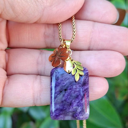 Collier en charoïte rectangulaire violette avec breloque feuille et fleur d'hibiscus dorées.
 dans la main
