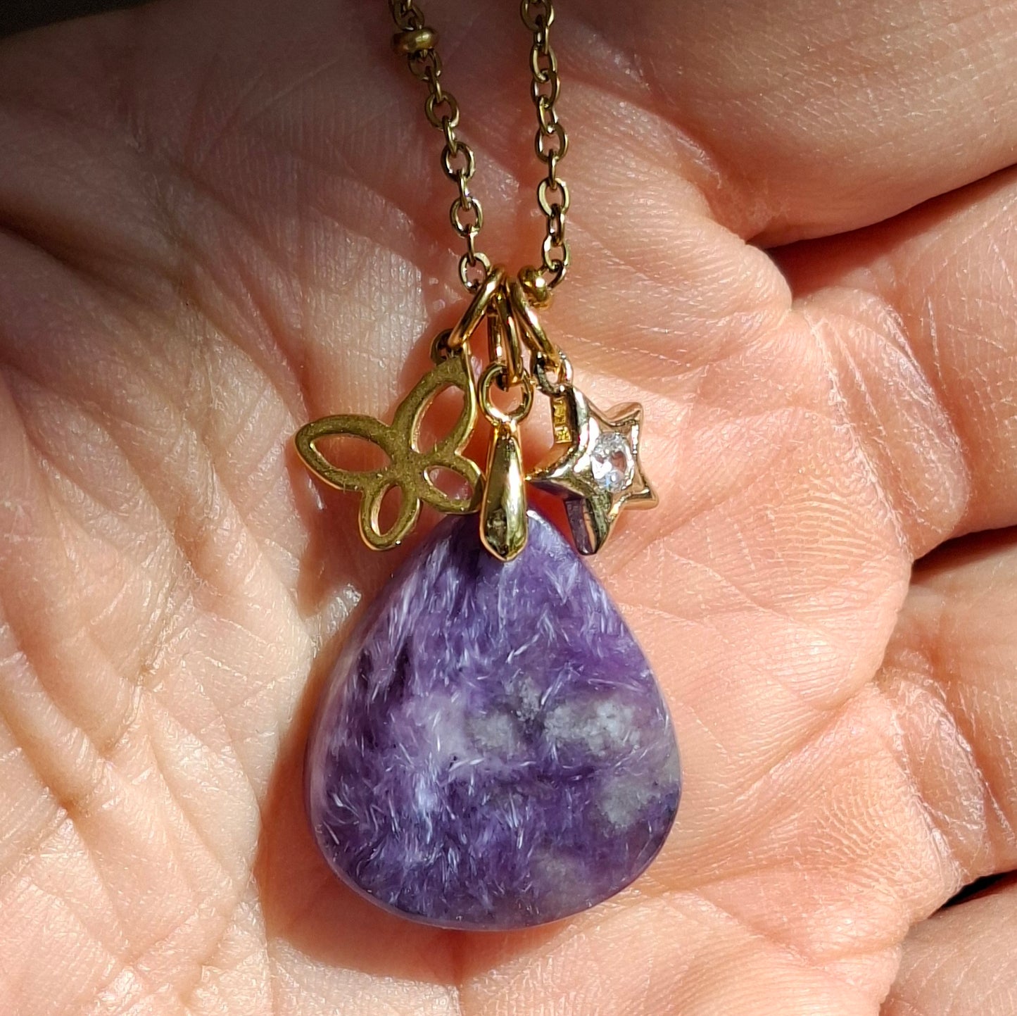 Collier en charoïte naturelle violette présenté dans la main, chaîne maille perlée acier inoxydable doré.
