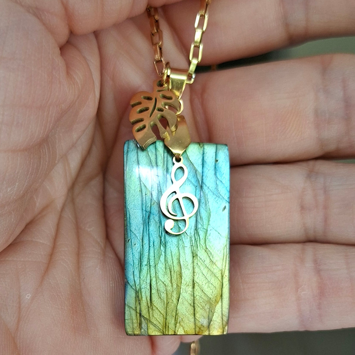 Collier acier doré en labradorite rectangulaire verte et dorée avec breloques feuille et clé de sol dans la main.