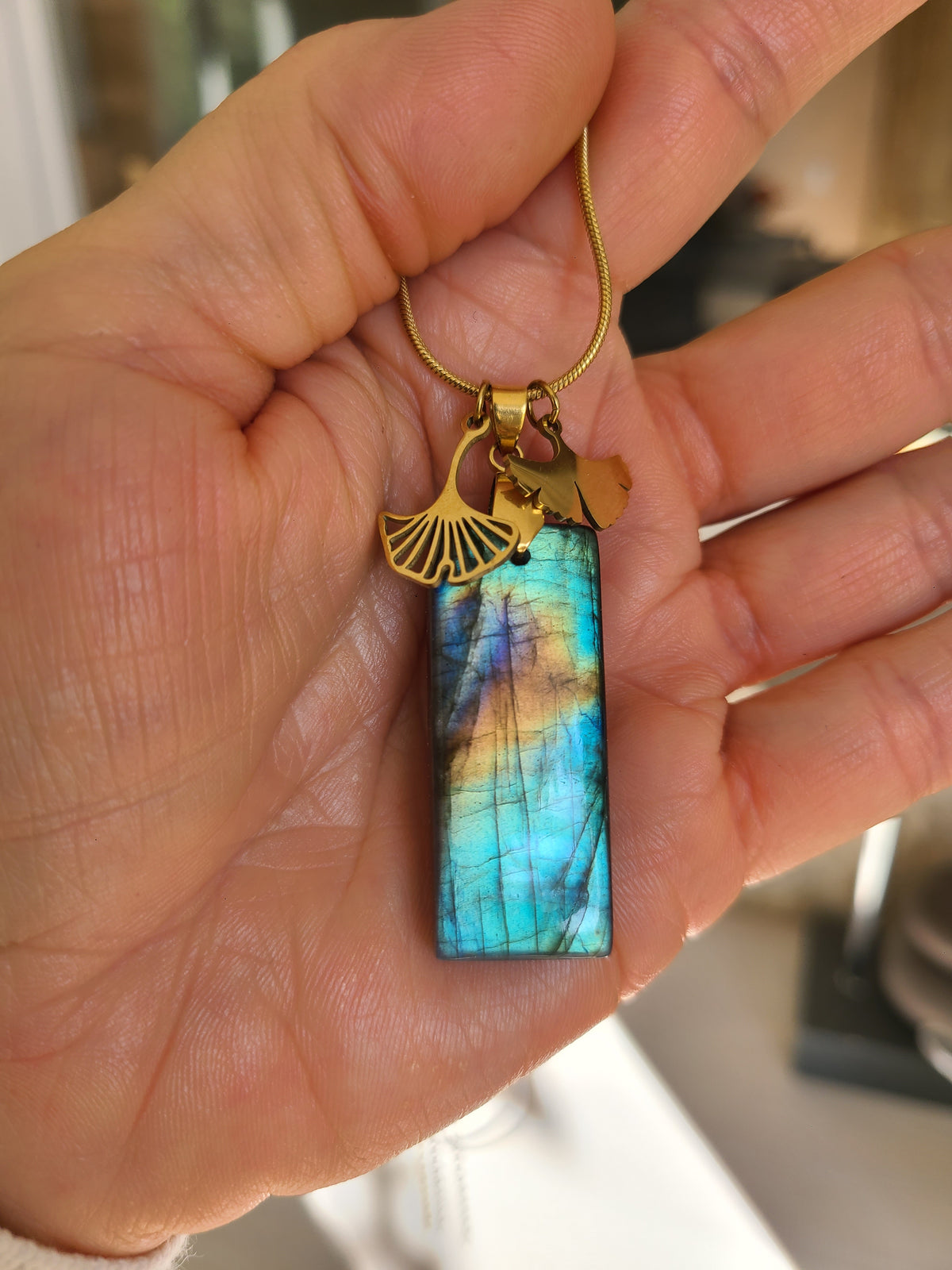 Collier acier doré en labradorite rectangulaire aux reflets multicolores avec breloques feuille de ginkgo dans une main. Petite Rainette Bijoux