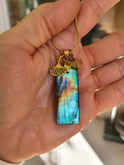 Collier acier doré en labradorite rectangulaire aux reflets multicolores avec breloques feuille de ginkgo dans une main. Petite Rainette Bijoux