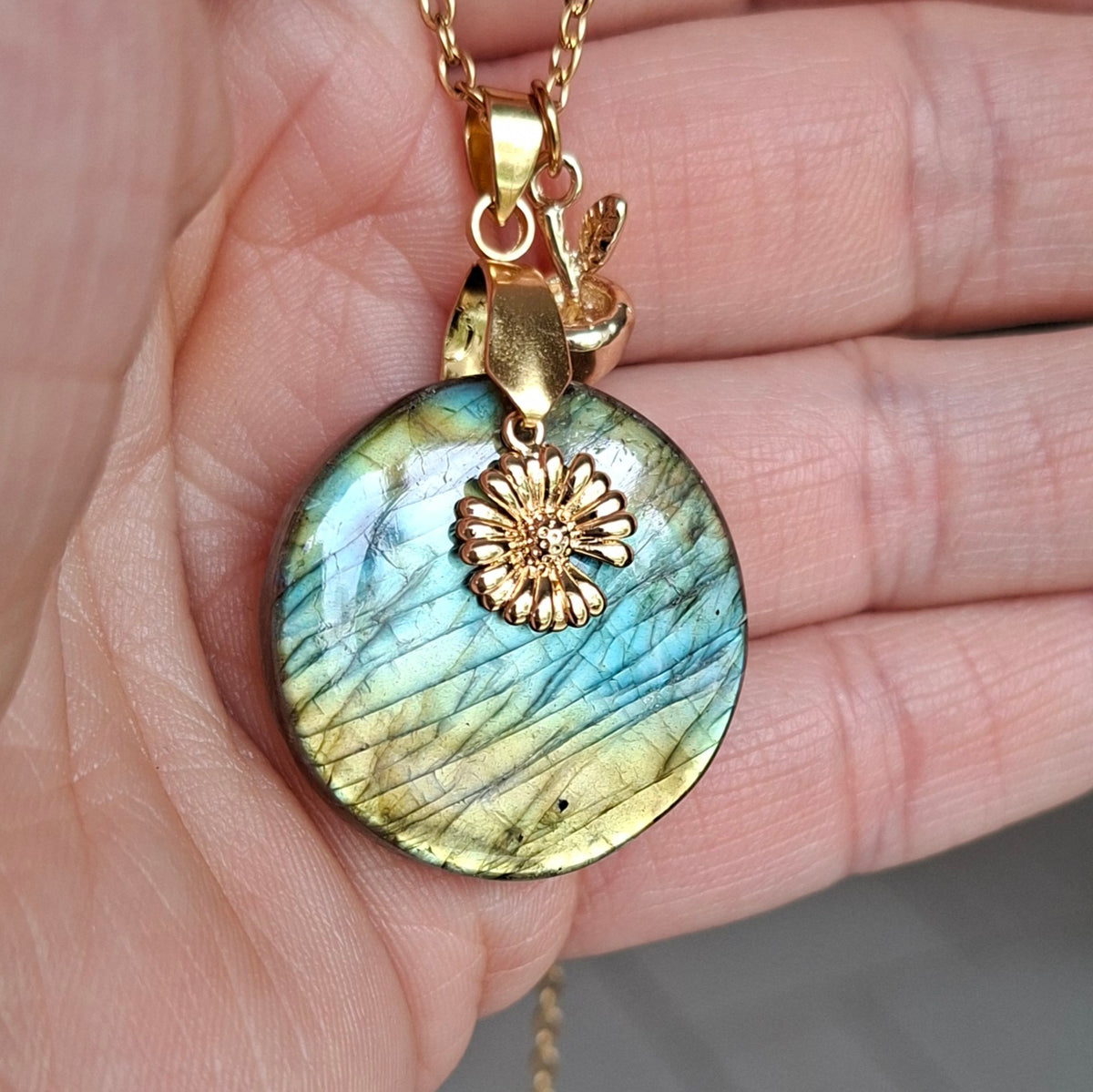 Collier acier doré en labradorite ronde présenté dans la main avec breloques fleur et pomme dorée.