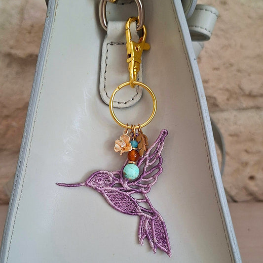 Bijou de sac Colibri Joie de vivre mauve porté sur un sac avec pierres naturelles et détails dorés