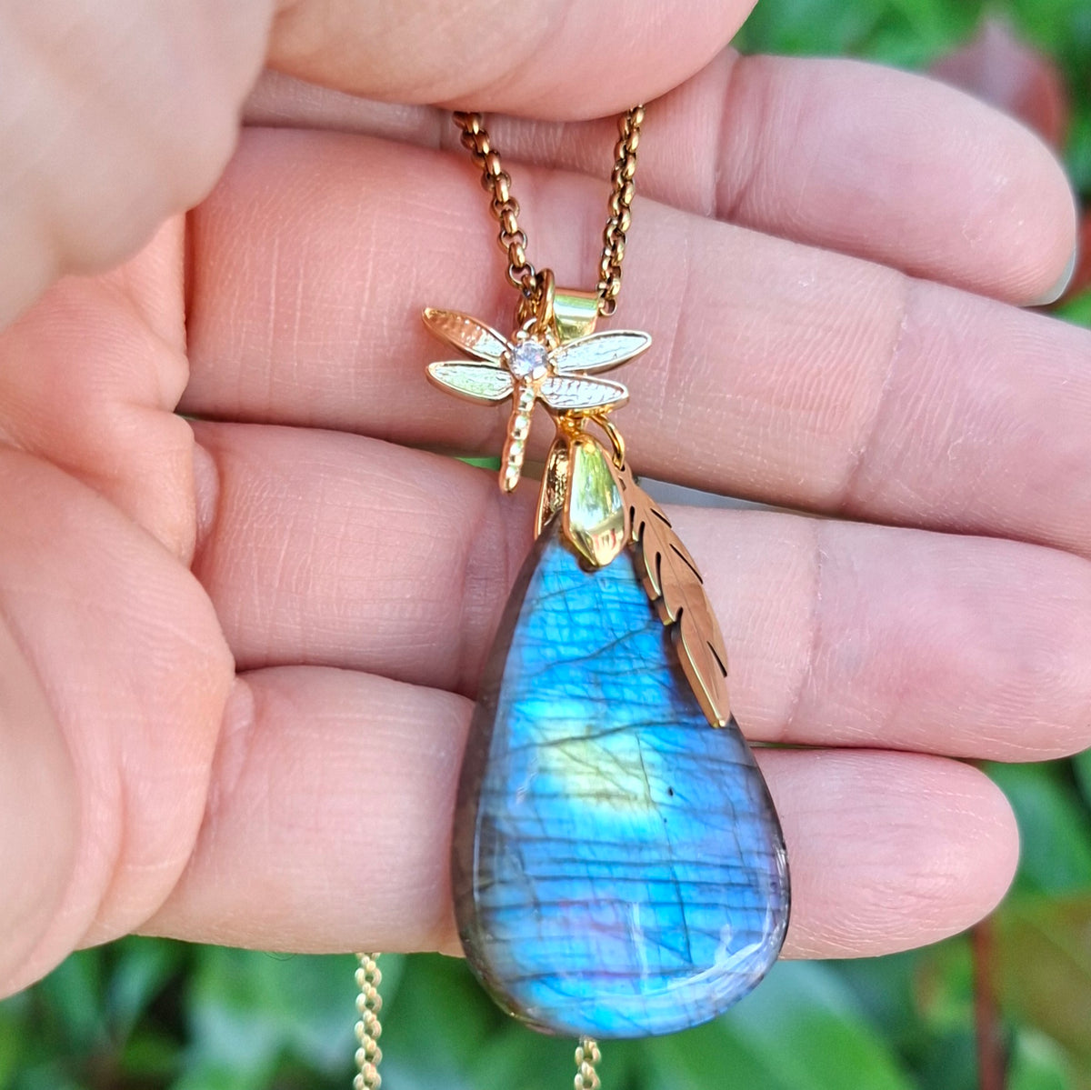 Collier en labradorite goutte bleue et dorée tenu dans la main avec breloque plume et libellule.