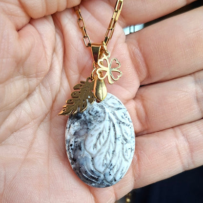 Collier en opale dendrite ciselée motif feuille présenté dans la main avec breloques végétales dorées.